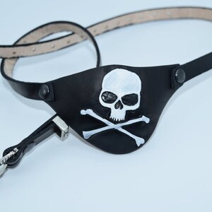 Op de afbeelding: Een zwart leren piratenooglap met een wit schedel- en kruisbeenderontwerp. De ooglap is bevestigd aan een zwart leren band met een zilveren gesp.