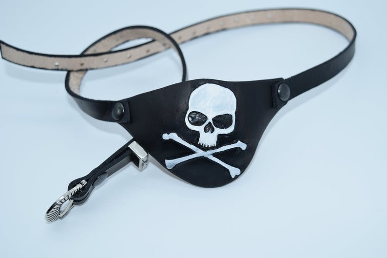 Op de afbeelding: Een zwart leren piratenooglap met een wit schedel- en kruisbeenderontwerp. De ooglap is bevestigd aan een zwart leren band met een zilveren gesp.