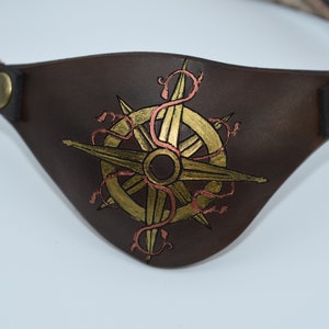 May include: A brown leather eye patch with a gold compass design and red vines. The compass has a gold needle and a gold circle around it.