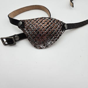 May include: A black leather eye patch with a silver and brown snakeskin pattern. The patch is attached to a black leather strap with a buckle.