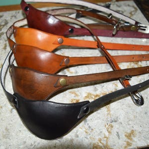 May include: Five leather eye patches in different colors, including brown, black, tan, and red. The eye patches are arranged in a stack, with the black eye patch on the bottom and the red eye patch on the top.