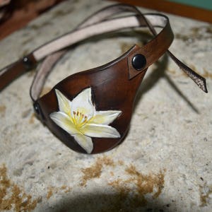 Op de afbeelding: Een bruin leren ooglapje met een wit bloemenontwerp. Het ooglapje heeft een band die om het hoofd gaat.