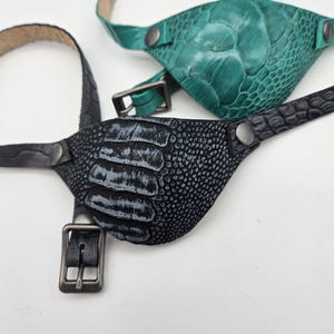 May include: Three leather eye patches in teal, black, and green, each with a textured, reptile-like pattern. The patches have adjustable straps with metal buckles and are secured with metal snaps. The background is plain white.