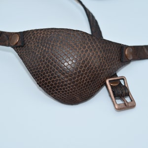 May include: A brown leather eye patch with a textured surface and a copper buckle. The patch is attached to a brown leather strap.