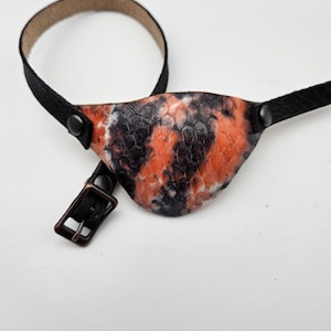 Può includere: Un paraluce per occhio in pelle nera con un motivo tie-dye arancione e nero. Il paraluce ha un cinturino regolabile con una fibbia.