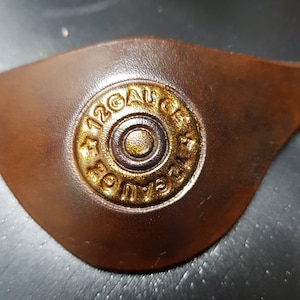 K&ouml;nnte beinhalten: Nahaufnahme eines braunen Lederflecks mit einem goldfarbenen 12 Gauge-Schrotflintenpatronenh&uuml;lse, die darauf gepr&auml;gt ist.