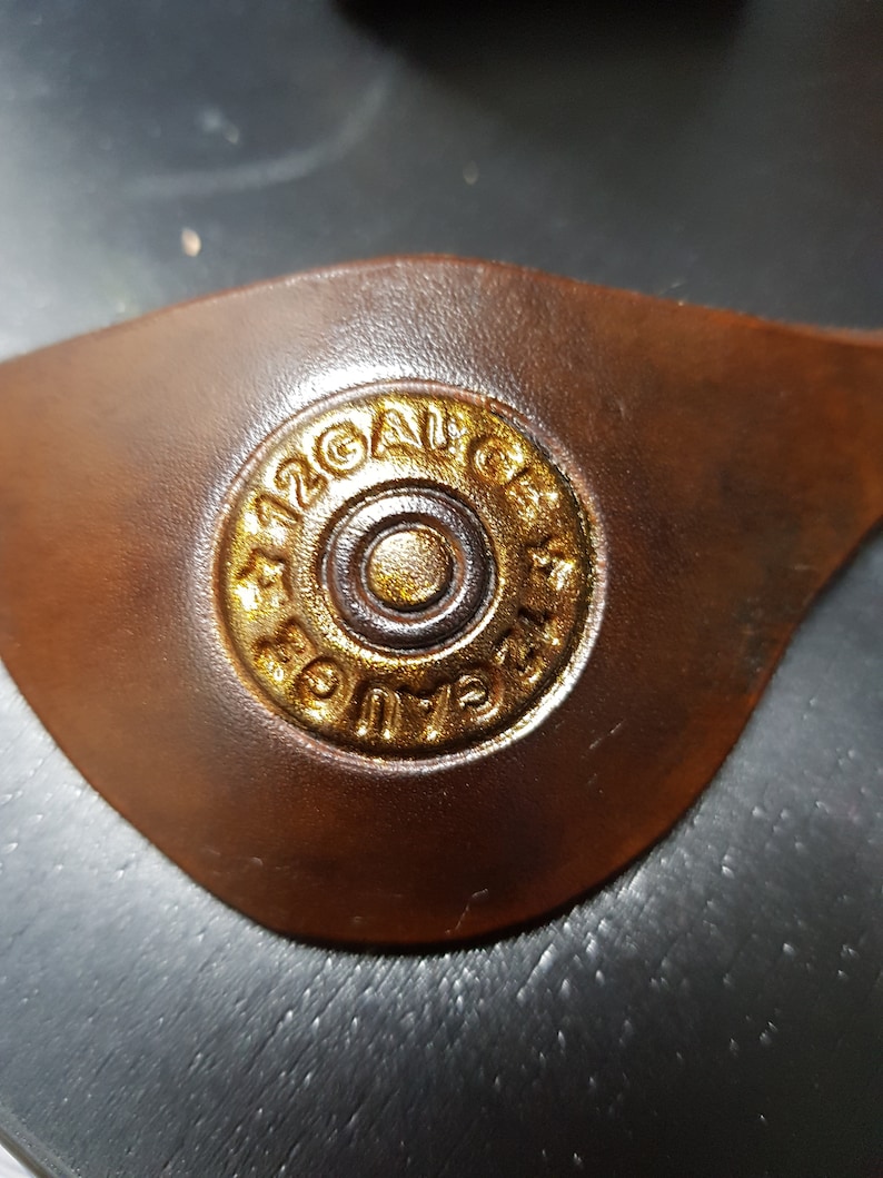 K&ouml;nnte beinhalten: Nahaufnahme eines braunen Lederflecks mit einem goldfarbenen 12 Gauge-Schrotflintenpatronenh&uuml;lse, die darauf gepr&auml;gt ist.