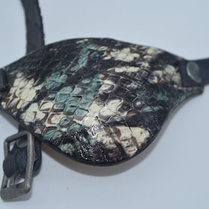 May include: A black leather eye patch with a green and white snakeskin pattern. The patch has a buckle strap and a metal eyelet.