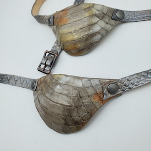 May include: Two handmade eye patches made of textured, grey-toned leather. The patches have a reptile-like pattern with hints of gold and orange. Each patch has a strap and a buckle or button closure. The background is white.