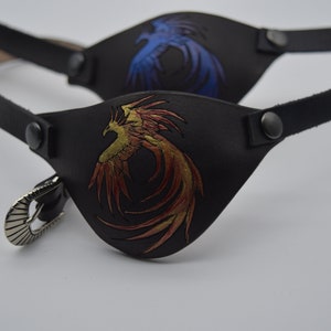 May include: Two black leather eye patches with adjustable straps. One patch features a gold and red phoenix design, while the other features a blue phoenix design.
