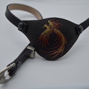 Puede incluir: Un parche para el ojo de cuero negro con un dise&ntilde;o de f&eacute;nix dorado y rojo. El parche para el ojo est&aacute; unido a una correa de cuero negro con una hebilla plateada.