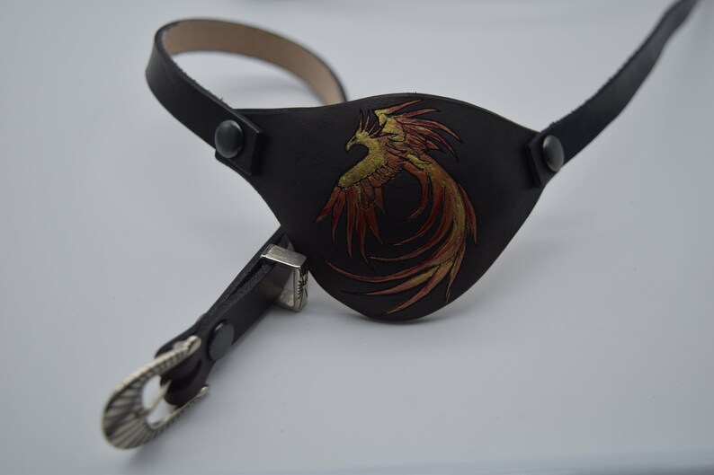 Puede incluir: Un parche para el ojo de cuero negro con un dise&ntilde;o de f&eacute;nix dorado y rojo. El parche para el ojo est&aacute; unido a una correa de cuero negro con una hebilla plateada.