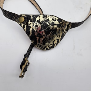 May include: Black and gold eye patch with a textured pattern. The eye patch has a strap and buckle for adjustment. The design features a gold metallic pattern on a black background. The strap is a light brown color.