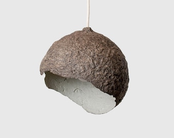 May include: A brown paper lampshade with a white interior, hanging from a white cord. The lampshade is made of recycled paper and has a unique, organic shape.
