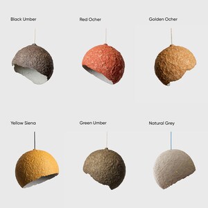 May include: Six different colored paper lampshades hanging from the ceiling. The colors are black umber, red ochre, golden ochre, yellow sienna, green umber, and natural grey.