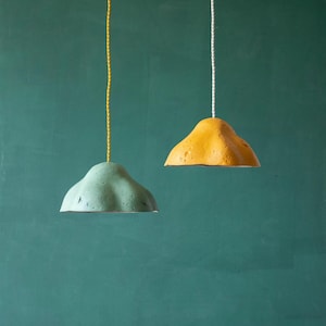 May include: Two pendant lights with a textured, cloud-like design. One is a muted teal color, the other is a mustard yellow. Each hangs from a cord, one yellow and one white, against a teal-green background. These lights are ideal for home decor.