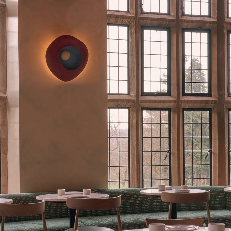 May include: A modern dining area with a large window overlooking a green landscape. The room features a long, green bench with a white marble table and chairs. A red and blue wall sconce is mounted on the wall.