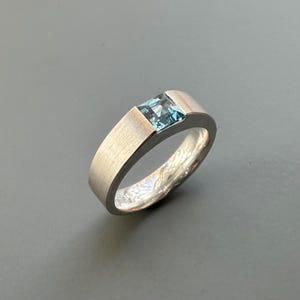 Op de afbeelding: Een zilveren ring met een lichtblauwe vierkante edelsteen in het midden. De ring heeft een geborstelde afwerking en een brede band.