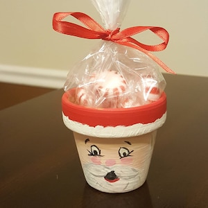 May include: A small terracotta pot painted to look like Santa Claus, filled with red and white peppermint candies and wrapped with a red ribbon and clear cellophane.