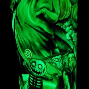 K&ouml;nnte beinhalten: Ein gr&uuml;ner, im Dunkeln leuchtender Becher mit einem Motiv von Figuren aus Nightmare Before Christmas. Die Figuren sind Jack Skellington, Sally und Oogie Boogie.