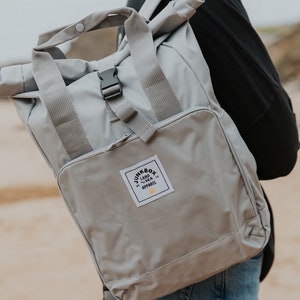 May include: A grey roll-top backpack with adjustable straps and a black buckle. The backpack has a white patch with the text "Junkbox Land & Sea Apparel" and a yellow sun graphic.