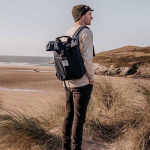 Könnte beinhalten: Eine Person mit einer schwarzen Mütze und einem beigefarbenen Langarmshirt mit einem dunkelblauen Rucksack steht auf einem Sandstrand mit hohem Gras.