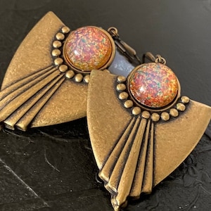 May include: A pair of Art Deco-style earrings. Each earring features a fan-shaped, bronze-colored metal base with a central, round, iridescent stone. The earrings have a lever-back closure and are set against a dark background.
