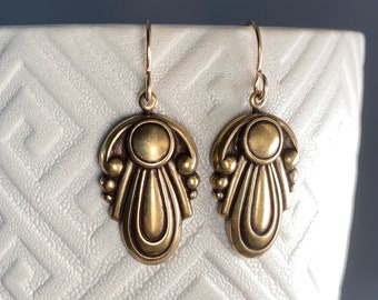 May include: A pair of gold-toned dangle earrings with a geometric design. The earrings feature a teardrop shape with a rounded top and a series of small circles around the edge.