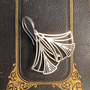 May include: A silver-colored earring with a fan-like design, featuring multiple curved lines and a hook closure. The earring is displayed on a textured black surface with gold decorative accents.