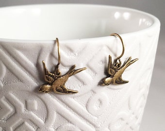 May include: A pair of gold-toned earrings featuring a delicate bird design. The earrings are hanging from a white ceramic bowl.