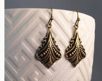 May include: A pair of gold-tone dangle earrings with a detailed, ornate design. The earrings have a teardrop shape with a pointed bottom and a series of small, round beads along the edges.
