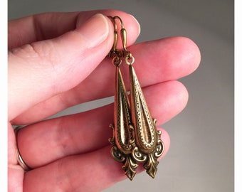 May include: A pair of antique-style brass earrings. The earrings have a long, teardrop shape with intricate filigree and decorative elements. The earrings are held up by a lever back closure.