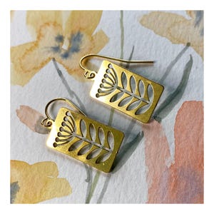 May include: A pair of gold-toned earrings with a delicate floral design. The earrings are rectangular in shape and feature a cutout design of a stylized flower with a stem and leaves.