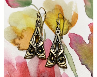 May include: A pair of gold-toned dangle earrings with a geometric design. The earrings are shaped like a triangle with a decorative pattern and a small flower at the top.
