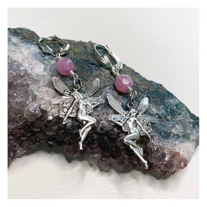 May include: A pair of silver earrings featuring a fairy design with pink gemstone accents. The fairies are depicted with wings and flowing hair.
