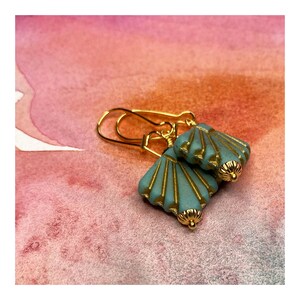 May include: A pair of gold earrings with turquoise blue shell-shaped charms. The charms have a gold-toned design and are attached to gold hooks.