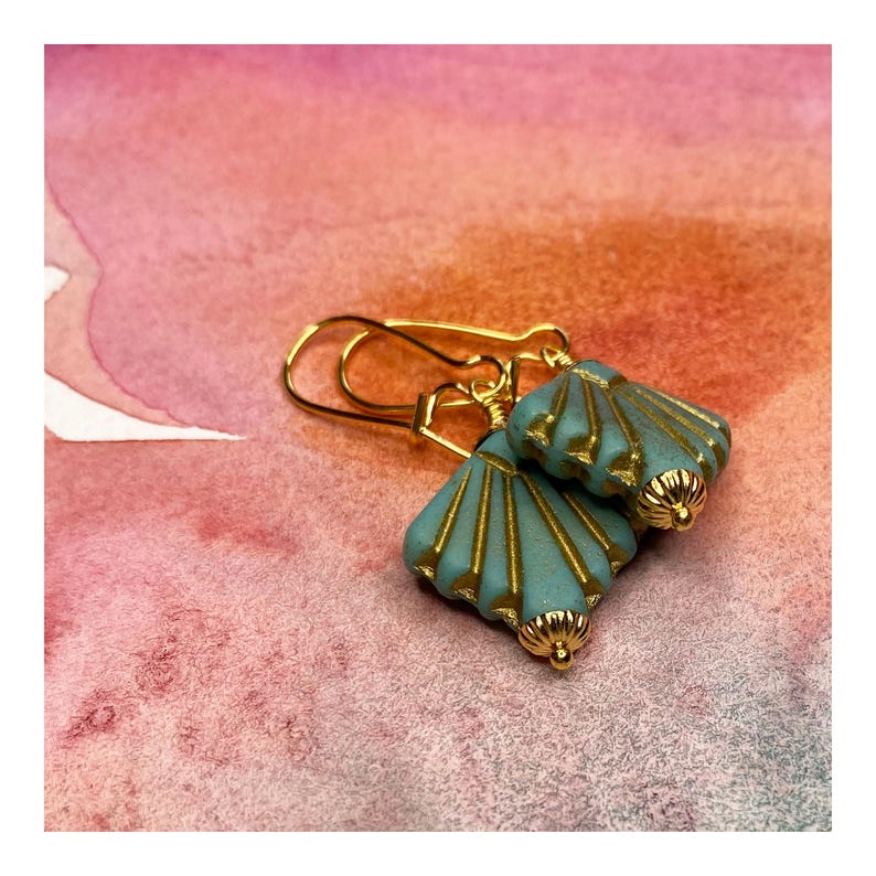 May include: A pair of gold earrings with turquoise blue shell-shaped charms. The charms have a gold-toned design and are attached to gold hooks.