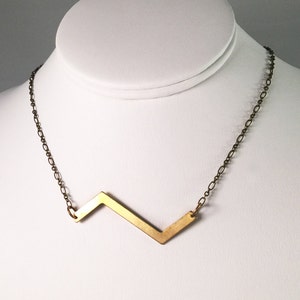 May include: A gold necklace with a geometric pendant shaped like a lightning bolt. The chain is a simple, delicate chain.