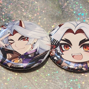 May include: Two round, colorful anime-style character buttons. One button features a character winking and giving a peace sign. The other button shows a character with a smiling face. Both buttons have white hair and red eyes.