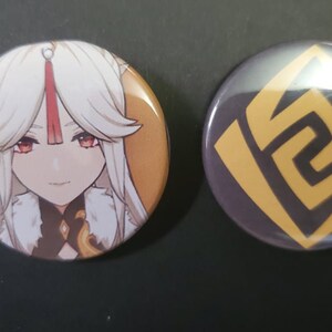 May include: Two round button pins. One features a cartoon anime character with white hair and red eyes. The other pin has a gold geometric design on a dark gray background.