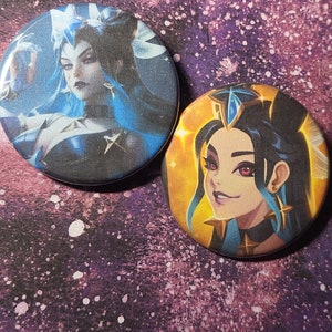 May include: Two round pinback buttons featuring a woman with blue hair and a crown. The first button has a dark blue background with stars and the woman is wearing a black and white outfit. The second button has a yellow background with the woman wearing a black and gold outfit.