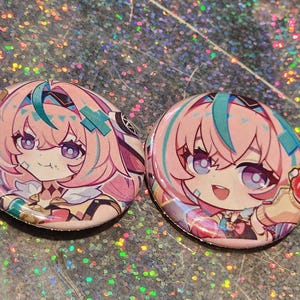 May include: Two round buttons featuring anime-style illustrations of a character with pink hair, teal accents, and purple eyes. The buttons have a glossy finish and are set against a sparkly background.
