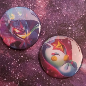 May include: Two round button pins with colorful images. The first pin features a purple and blue character with a yellow star on a dark background. The second pin features a white character with a red star on a blue background.