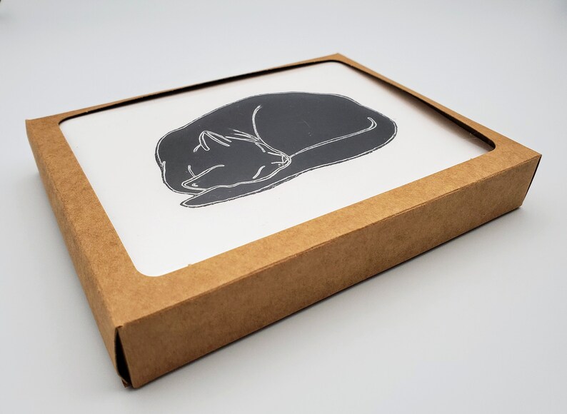 May include: A brown cardboard box with a white card inside. The card features a black and white line drawing of a cat curled up asleep.