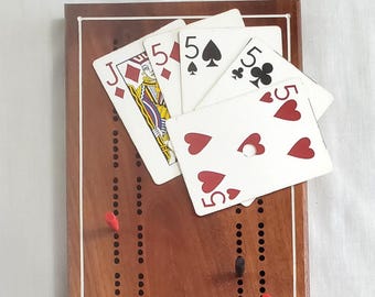 May include: A wooden cribbage board with a white border and the word "CRIBBAGE" printed in white. There are five playing cards on top of the board, including a Jack of Diamonds, a 5 of Diamonds, a 5 of Spades, a 5 of Clubs, and a 5 of Hearts.