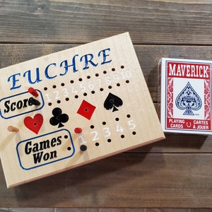 May include: A wooden Euchre scorekeeping board with red and black pegs, a red heart, a black spade, and a red diamond. The board has a white background with blue text that says "Euchre", "Score", "Games Won", and numbers 1 through 10. A deck of Maverick playing cards is in the background.