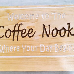 May include: Wooden sign with the words "Welcome to The Coffee Nook Where Your Day Begins" carved into the wood. The words "Coffee Nook" are in a dark brown color, and the other words are in white.