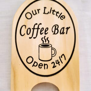 May include: Wooden sign with the text "Our Little Coffee Bar" in a black, circular frame. A steaming coffee mug graphic is centered below the text. The sign reads "Open 24/7" in black lettering. The sign has a light wood finish.