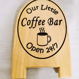 May include: Wooden sign with the text "Our Little Coffee Bar Open 24/7" in black lettering. A coffee mug graphic is in the center. The sign is light brown with a rounded top and a rectangular base with a notch.