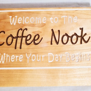 May include: Wooden sign with the words "Welcome to The Coffee Nook Where Your Day Begins" in white and brown lettering. The sign has a light brown background with a white border.